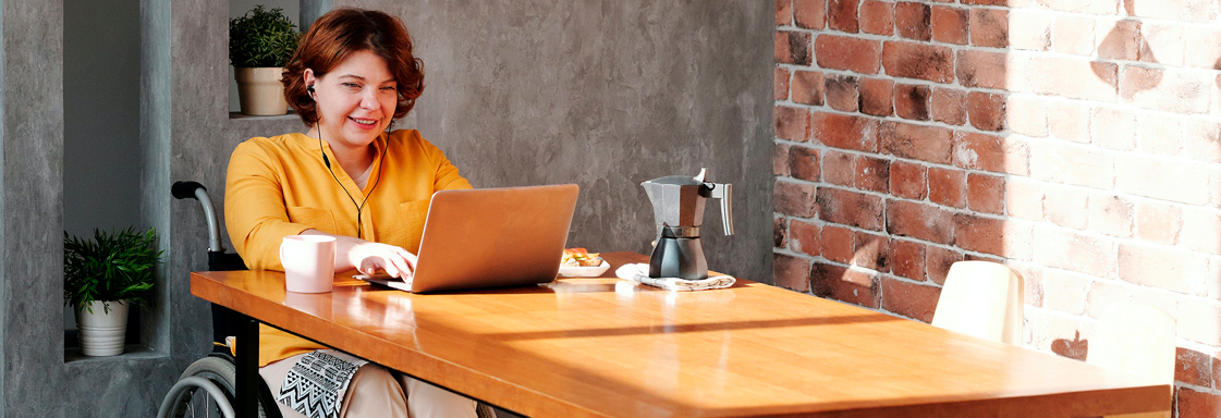Fotografía de mujer, usuaria de silla de ruedas, sentada frente a un escritorio, con una computadora portátil y una taza de café